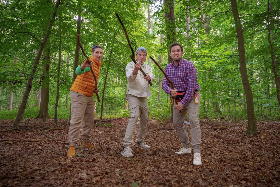 Auf dem Bild sieht man drei Personen, mitten in einem Wald. Sie haben einem Stock in der Hand und gucken böse nach vorne.